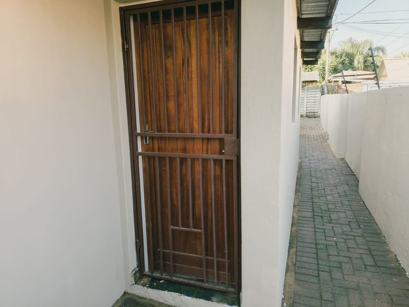 A side exterior view showing a wooden door with a security gate, leading to a paved walkway along the boundary wall.