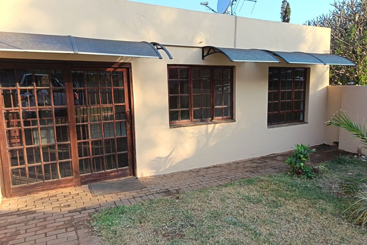 Rear exterior view of the apartment featuring timber-framed windows and sliding doors with protective polycarbonate awnings and a paved garden walkway.