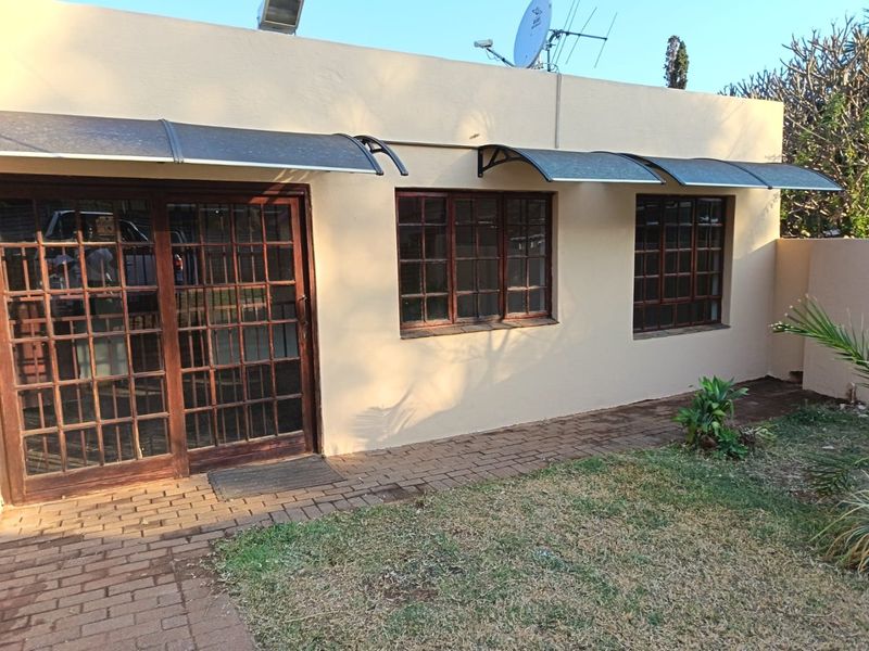 Rear exterior view of the apartment featuring timber-framed windows and sliding doors with protective polycarbonate awnings and a paved garden walkway.