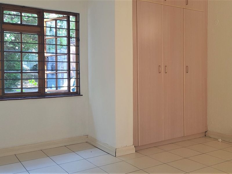 A bright, empty bedroom featuring tiled flooring, built-in cupboards, and timber-framed windows with security bars overlooking the garden.