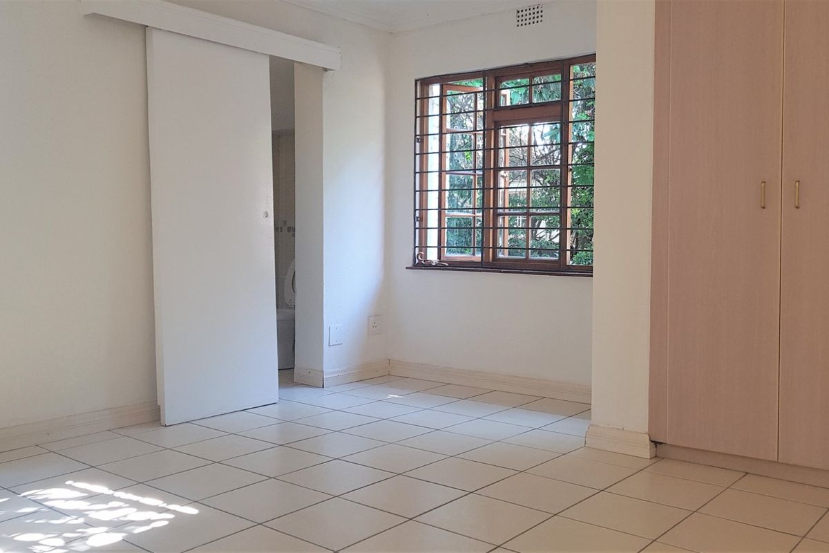A bright, empty bedroom featuring tiled flooring, built-in cupboards, and timber-framed windows with security bars, offering a clean and functional living space.