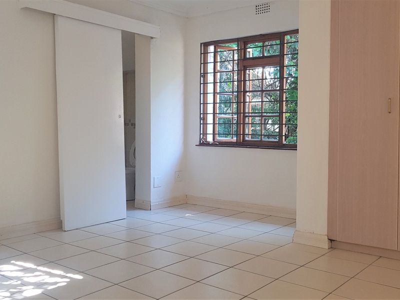 A bright, empty bedroom featuring tiled flooring, built-in cupboards, and timber-framed windows with security bars, offering a clean and functional living space.