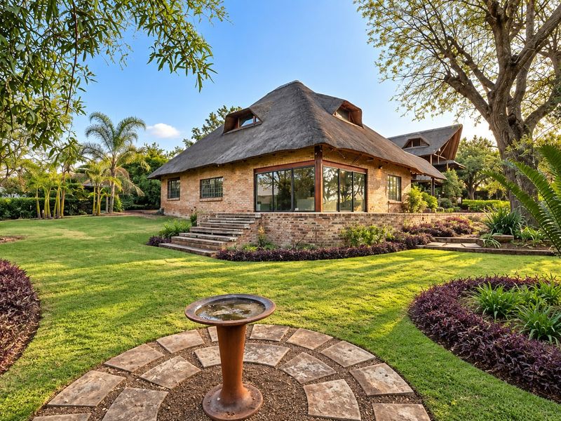 A well-maintained house with a traditional thatched roof, brick exterior, and expansive landscaped garden featuring stone pathways and mature trees.