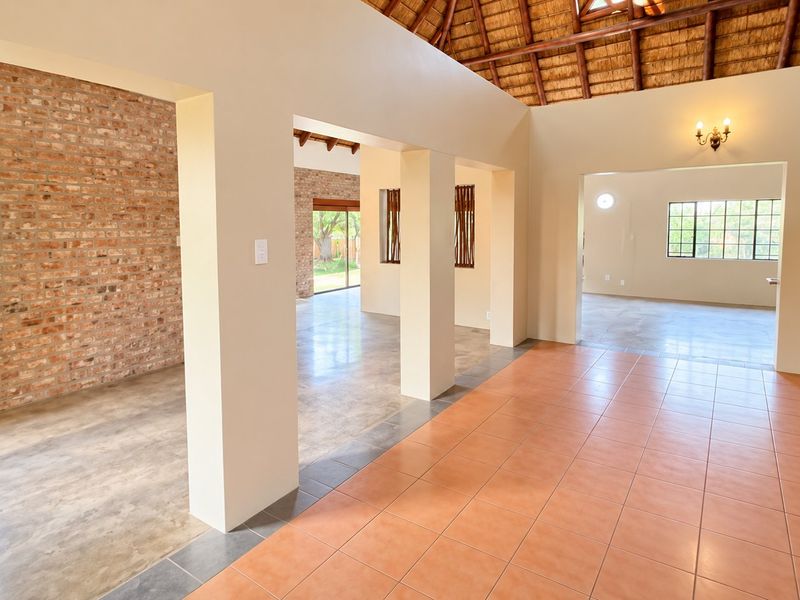 Spacious open-plan living area featuring a high thatched ceiling with exposed rafters, a mix of tiled and polished concrete floors, and an exposed brick wall.