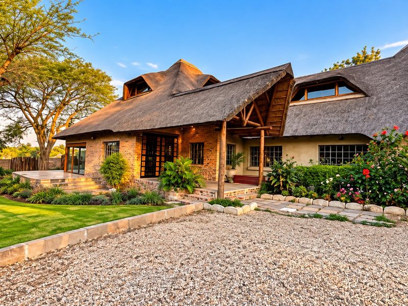 A charming, well-maintained house featuring a traditional thatch roof, exposed brick and stone facade, and a beautifully landscaped garden with a gravel driveway.