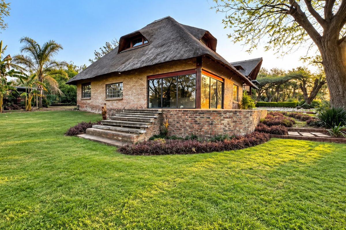 A charming rear view of a thatched-roof house featuring exposed brickwork, large glass sliding doors, and a well-maintained garden setting.