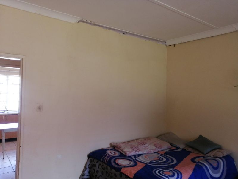 A bedroom featuring plain walls and visible ceiling damage near the cornice, with an adjacent view into a kitchen area.