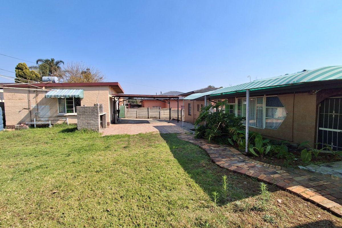 Rear exterior view featuring a paved patio area, built-in brick braai, covered carport, and a paved walkway leading through the garden.