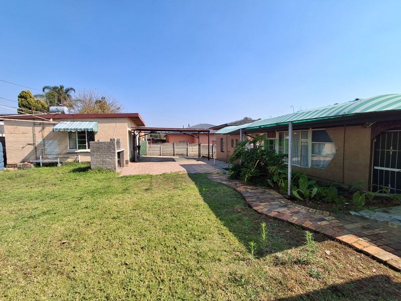 Rear exterior view featuring a paved patio area, built-in brick braai, covered carport, and a paved walkway leading through the garden.