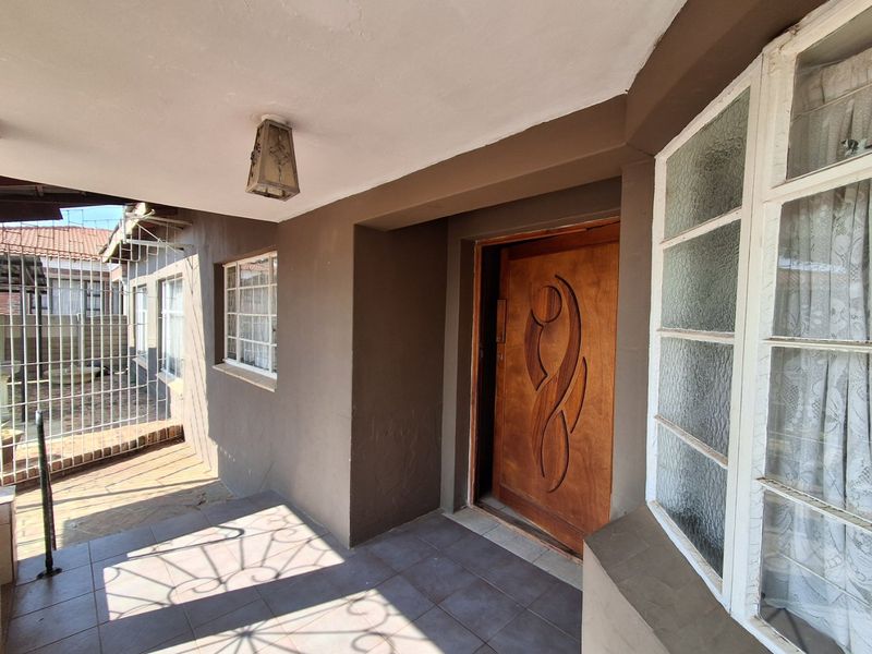 A covered entrance patio featuring tiled flooring, a decorative wooden front door, and steel security fixtures, providing a functional outdoor transition space.