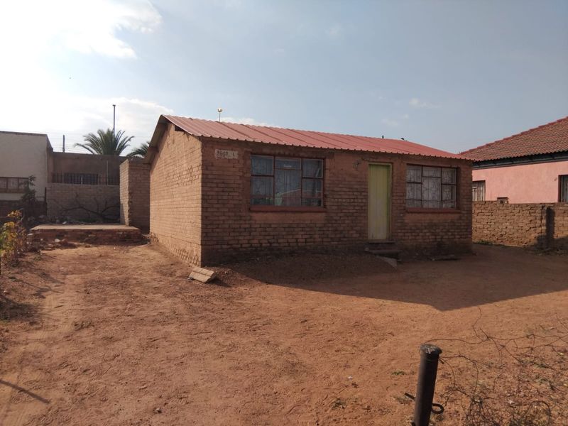 A single-story detached house featuring an exposed brick exterior, corrugated metal roof, and steel-framed windows, situated on an unpaved plot with perimeter walling.