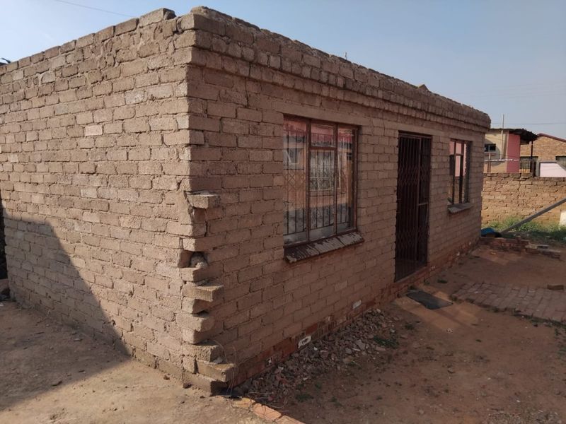 A single-story residential structure featuring unplastered brick walls, metal window frames, and a security gate, showing signs of structural wear at the corner.