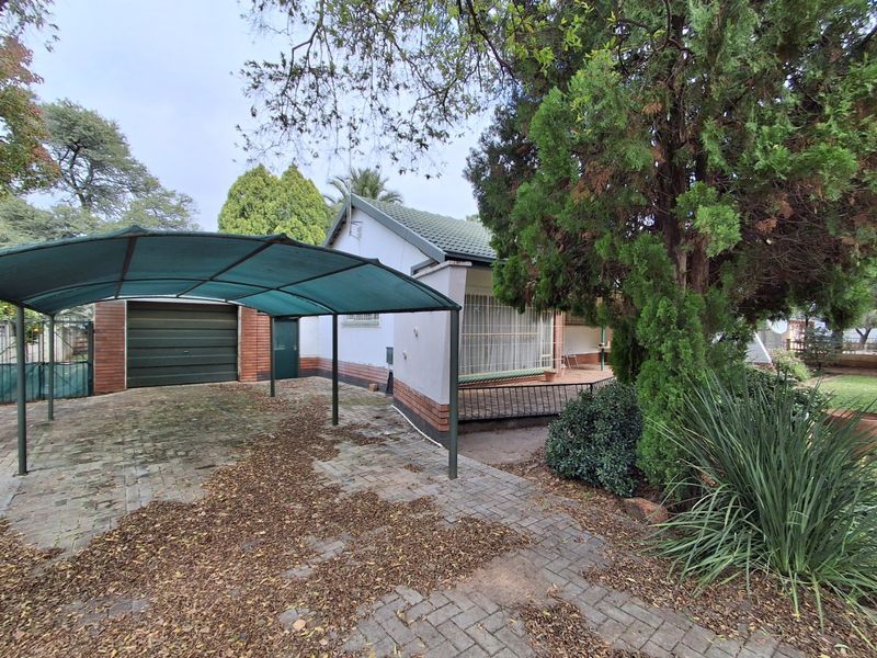 Front exterior view of the house featuring a paved driveway, a covered carport, and a single garage, surrounded by established garden trees.