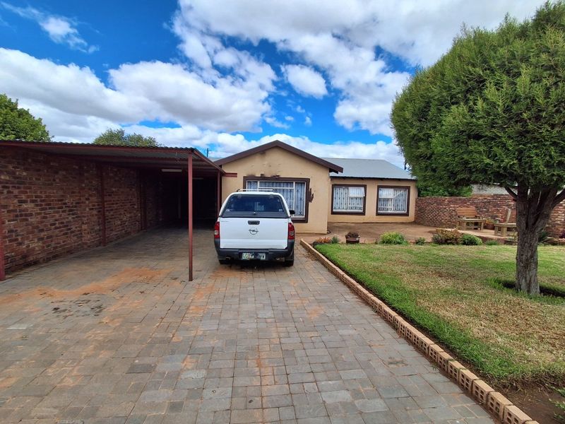 Front view of a single-story house featuring a paved driveway, attached carport, and a landscaped front garden with brick boundary walling.