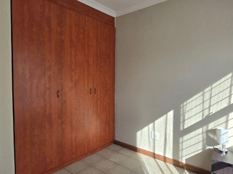 A bright, empty bedroom featuring built-in wooden cupboards, tiled flooring, and natural light streaming through the window.