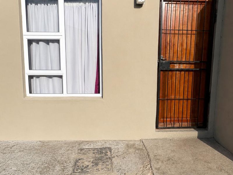 Exterior view of a residential entrance featuring a wooden door with a security gate, an aluminium window, and a concrete apron.