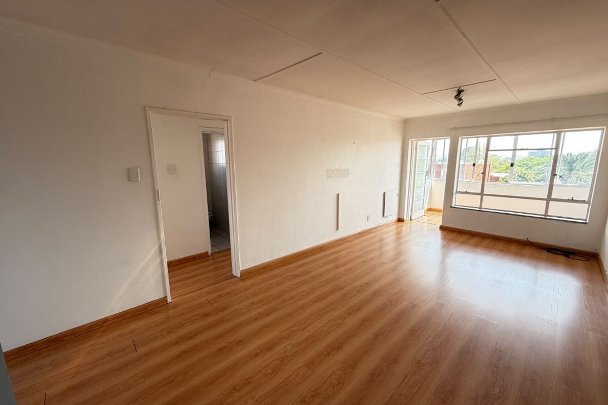 An empty, sunlit living room featuring laminate flooring, large aluminium windows, and direct access to an adjoining balcony.
