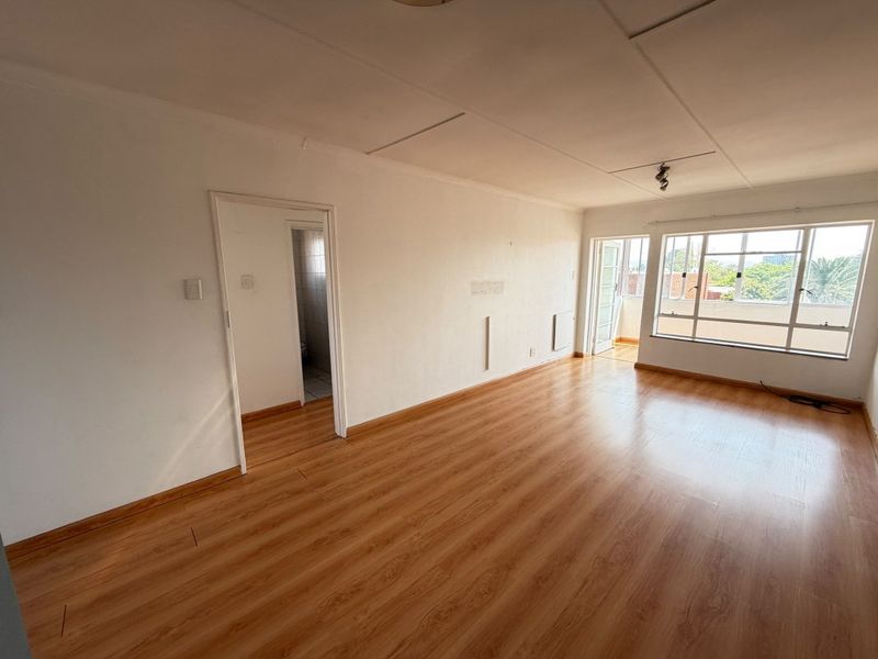 An empty, sunlit living room featuring laminate flooring, large aluminium windows, and direct access to an adjoining balcony.