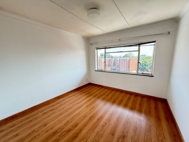 A bright, empty bedroom featuring laminate wood flooring, a large window with a curtain rod, and a simple ceiling light fixture.