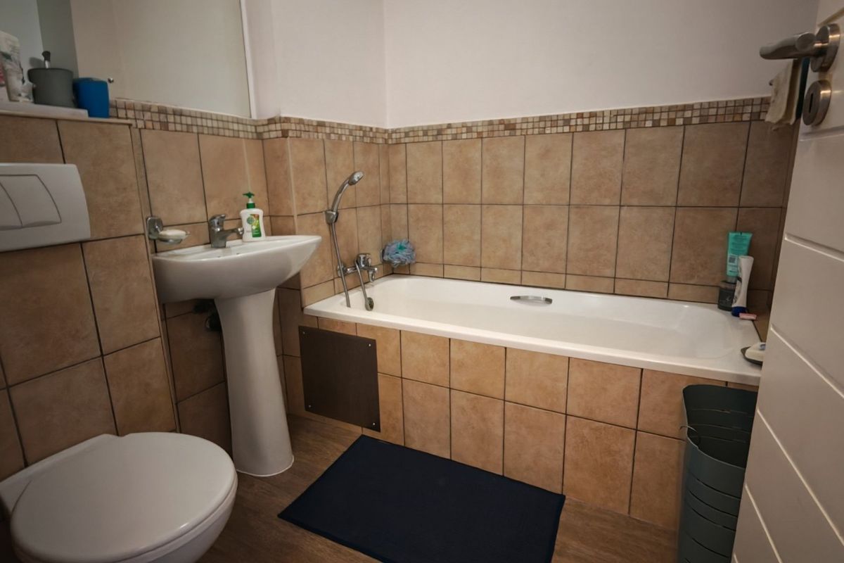 A functional bathroom featuring a pedestal basin, bathtub with shower mixer, and wall-mounted toilet, finished with neutral wall tiling.