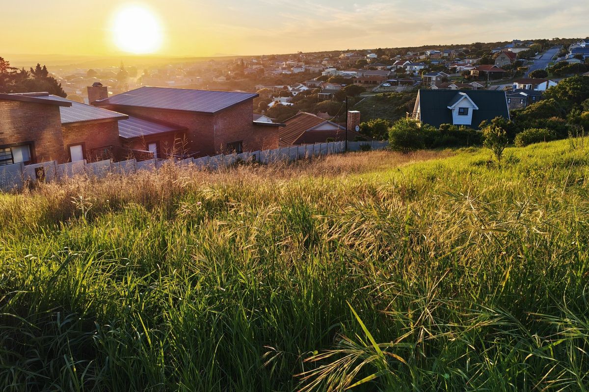 A scenic view overlooking a residential neighborhood from a sloped, undeveloped grassy plot at sunset.