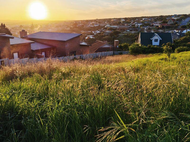 A scenic view overlooking a residential neighborhood from a sloped, undeveloped grassy plot at sunset.