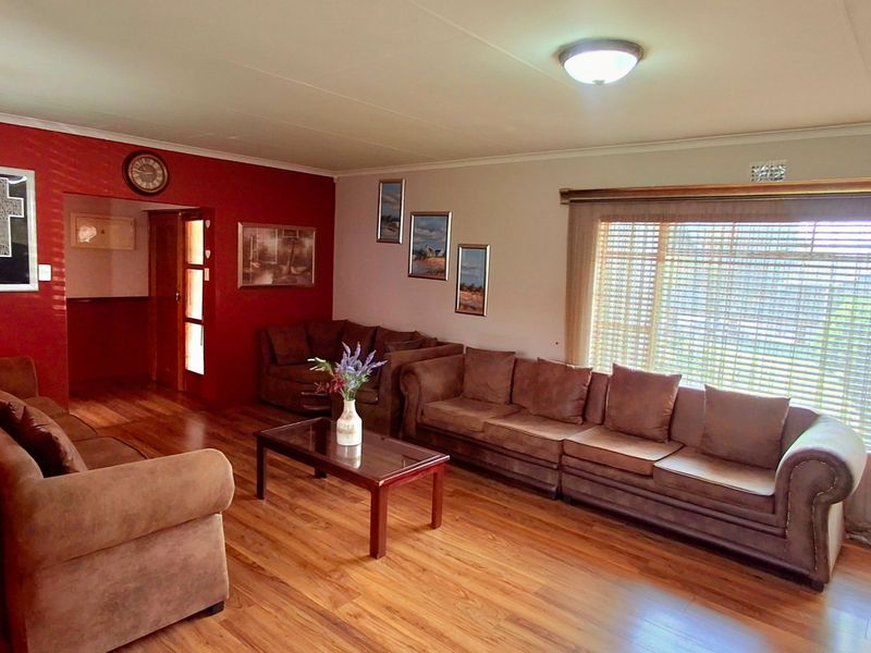 A spacious living room featuring laminate flooring, neutral walls with a red accent wall, and large windows fitted with venetian blinds.