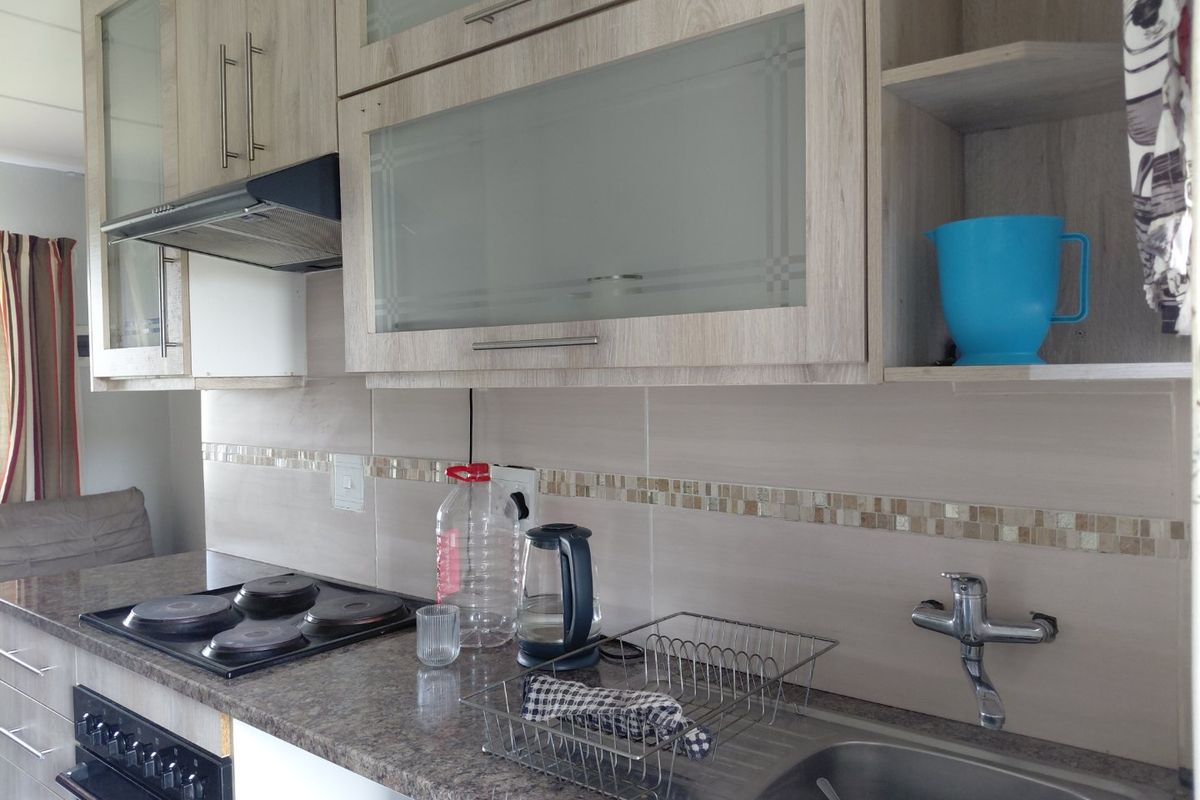 A functional kitchen featuring light wood-grain cabinetry, granite-look laminate countertops, a built-in electric hob and oven, and a stainless steel sink with tiled backsplash.