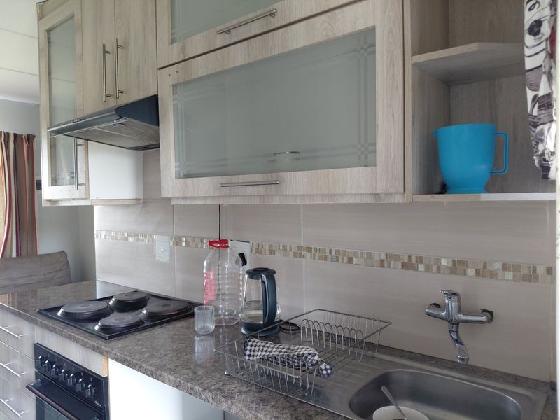 A functional kitchen featuring light wood-grain cabinetry, granite-look laminate countertops, a built-in electric hob and oven, and a stainless steel sink with tiled backsplash.