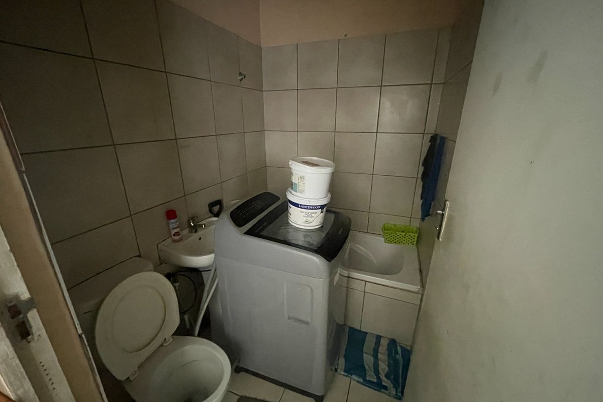 A compact bathroom featuring a toilet, small washbasin, and built-in bath with tiled finishes, currently utilized as a laundry area.