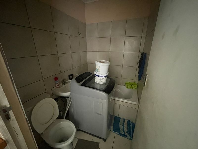 A compact bathroom featuring a toilet, small washbasin, and built-in bath with tiled finishes, currently utilized as a laundry area.