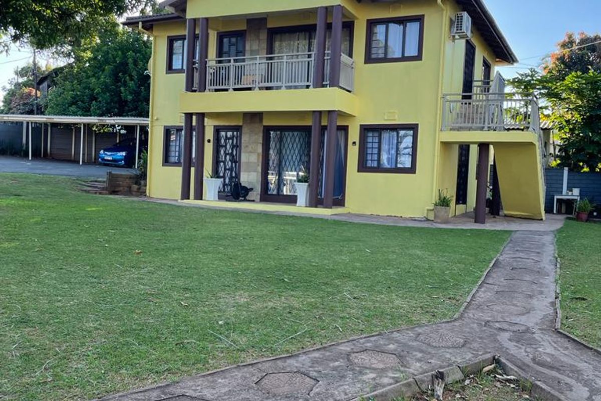 A two-story residential building featuring a yellow facade, upper-level balcony, external staircase, and a spacious surrounding lawn with a paved walkway.