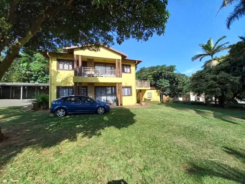 A two-story residential building featuring a yellow facade, upper-level balcony, and a spacious front lawn area.