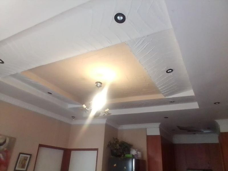 A view of a living room ceiling featuring a recessed design with integrated downlights and a central chandelier, showing signs of ceiling damage.