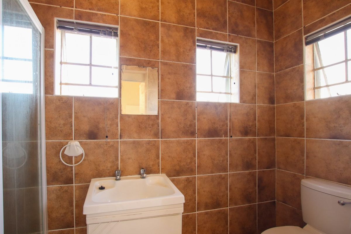 A functional bathroom featuring a shower enclosure, vanity basin, and toilet, finished with dated brown wall tiles and multiple windows for natural light.