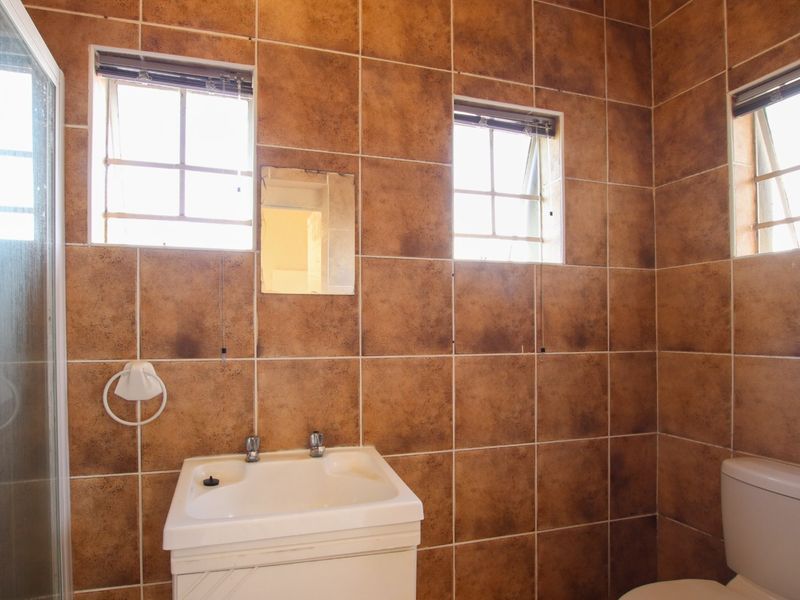 A functional bathroom featuring a shower enclosure, vanity basin, and toilet, finished with dated brown wall tiles and multiple windows for natural light.