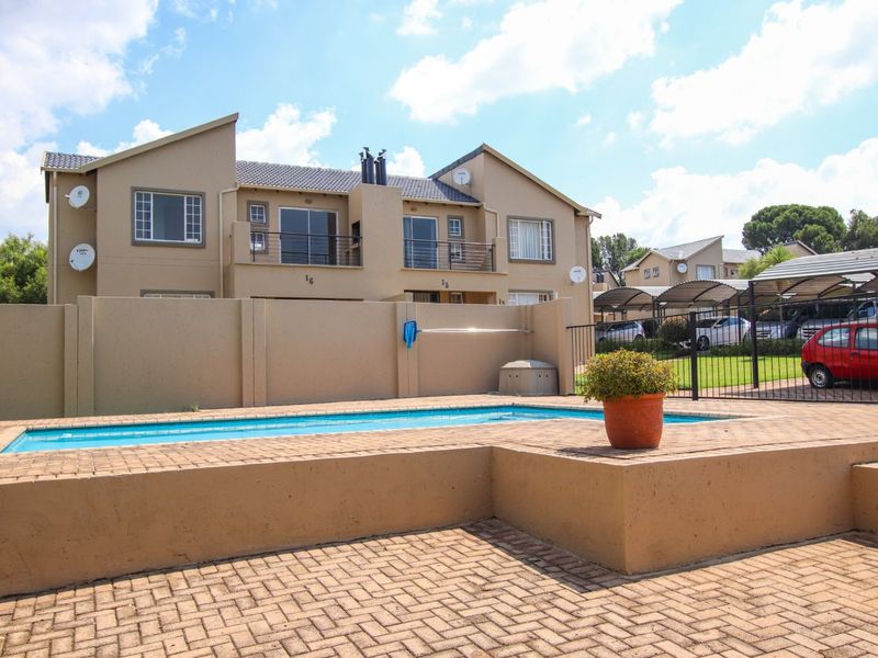 Exterior view of a residential apartment complex featuring a private swimming pool, paved deck area, and secure perimeter fencing with adjacent carport parking.