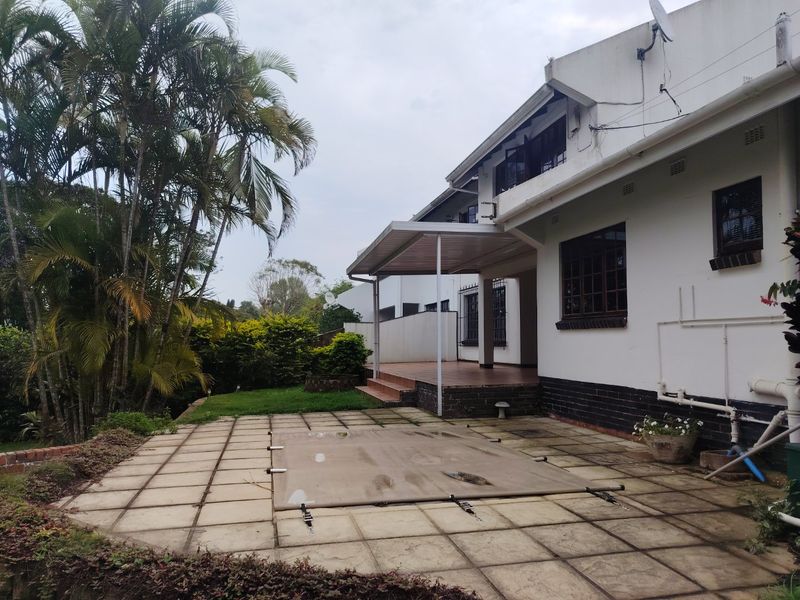 Rear exterior view featuring a covered patio area, paved pool deck with cover, and established garden surroundings.