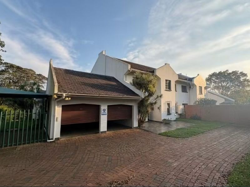 Front view of a residential property featuring a double garage, brick paved driveway, and a carport, set within a landscaped garden area.