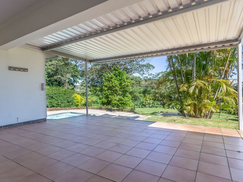 A spacious covered patio featuring tiled flooring, overlooking a private swimming pool and lush garden area, perfect for outdoor entertaining.