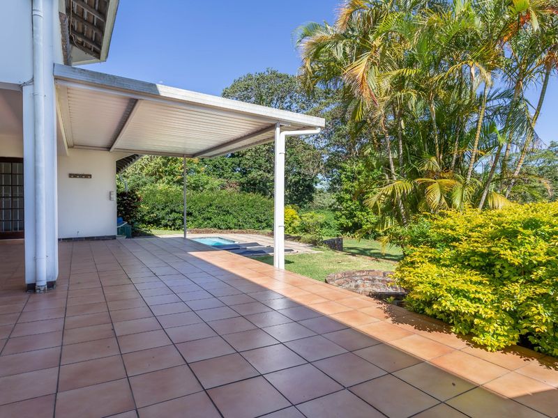 A spacious covered patio featuring tiled flooring, overlooking a private swimming pool and a lush, well-maintained garden area.