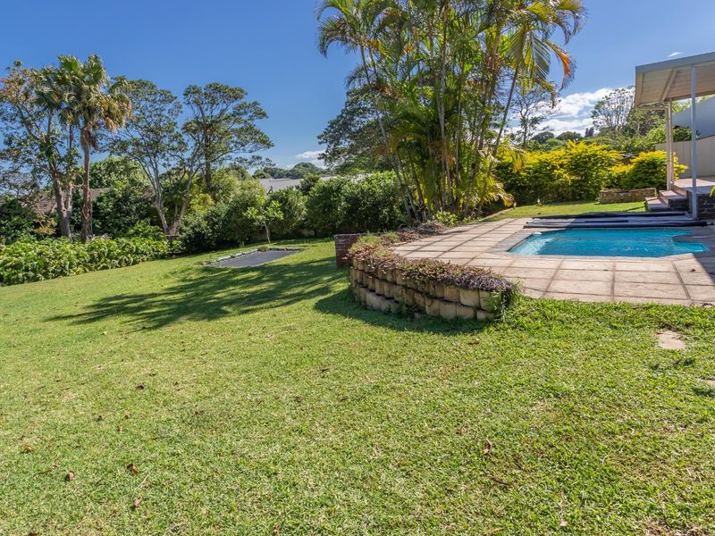 A spacious backyard featuring a swimming pool with a paved deck, a covered patio area, and a well-maintained lawn surrounded by mature trees.
