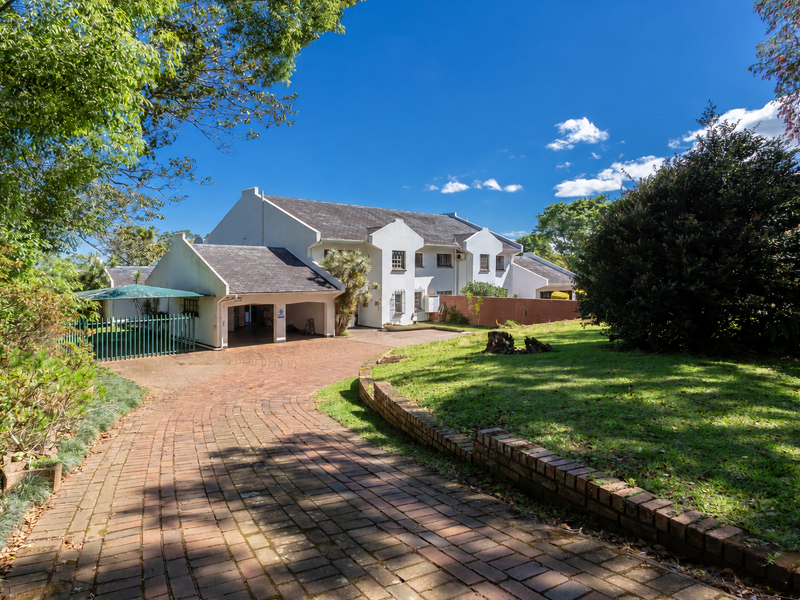 A view of the front elevation featuring a brick paved driveway leading to a covered carport, surrounded by a well-established garden.