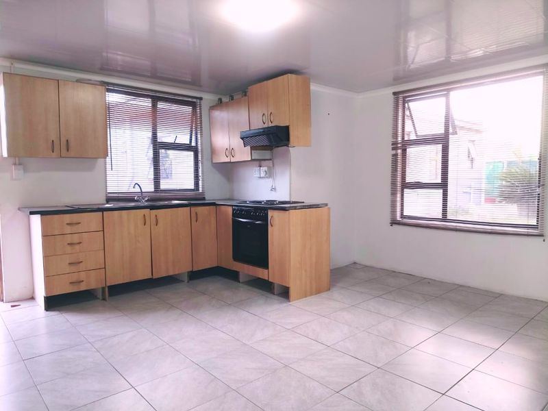 A functional, empty kitchen featuring light wood-tone cabinetry, built-in oven, extractor fan, and tiled flooring with ample natural light from two windows.