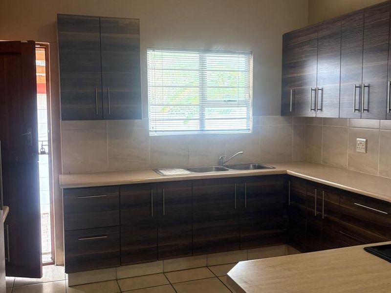 A functional kitchen featuring dark wood-look cabinetry, laminate countertops, a double sink, and neutral ceramic floor tiles with natural light from a window.