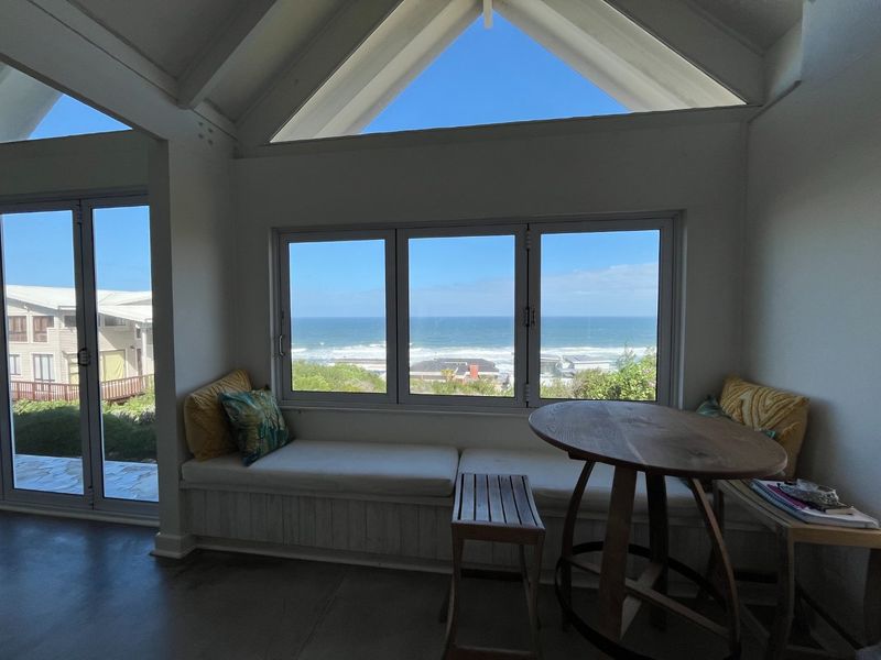 Bright living area featuring a vaulted ceiling, exposed rafters, and a built-in window seat offering expansive ocean views through large windows.