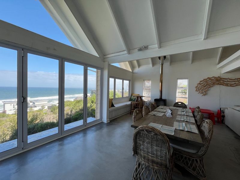 Spacious open-plan dining area featuring vaulted ceilings, polished concrete floors, a modern fireplace, and expansive glass doors offering panoramic ocean views.