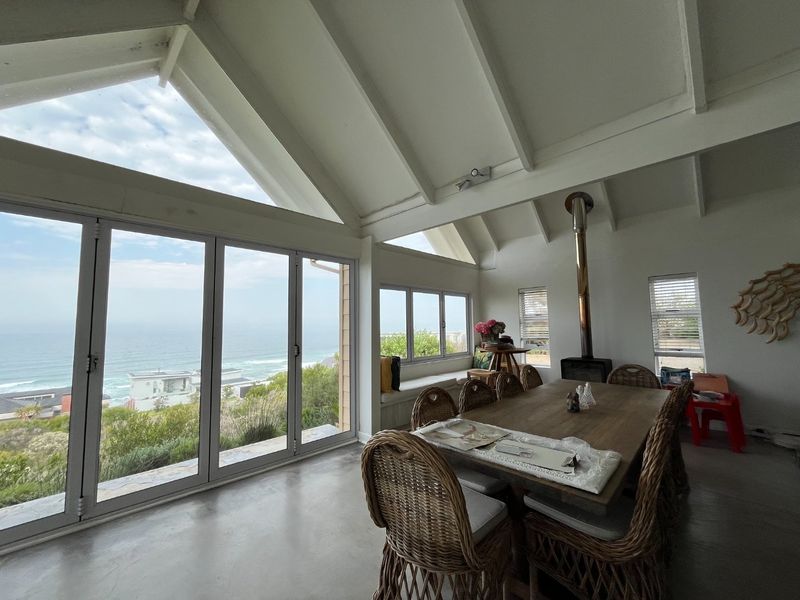Spacious dining area featuring a vaulted ceiling with exposed beams, polished concrete floors, a freestanding fireplace, and expansive bi-fold doors offering panoramic ocean views.