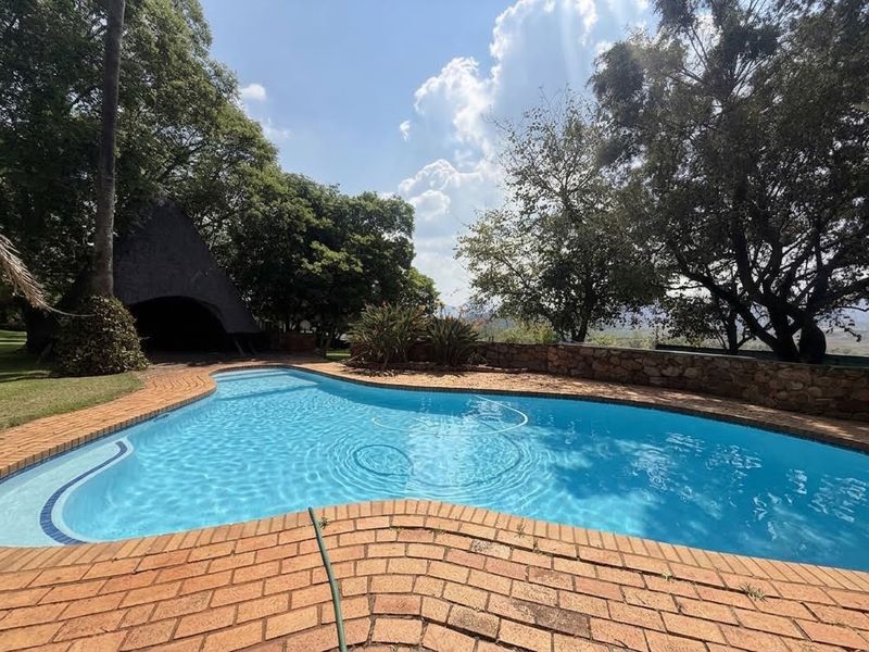 Outdoor swimming pool area featuring brick paving, a stone retaining wall, and a thatched lapa structure set within a mature garden.