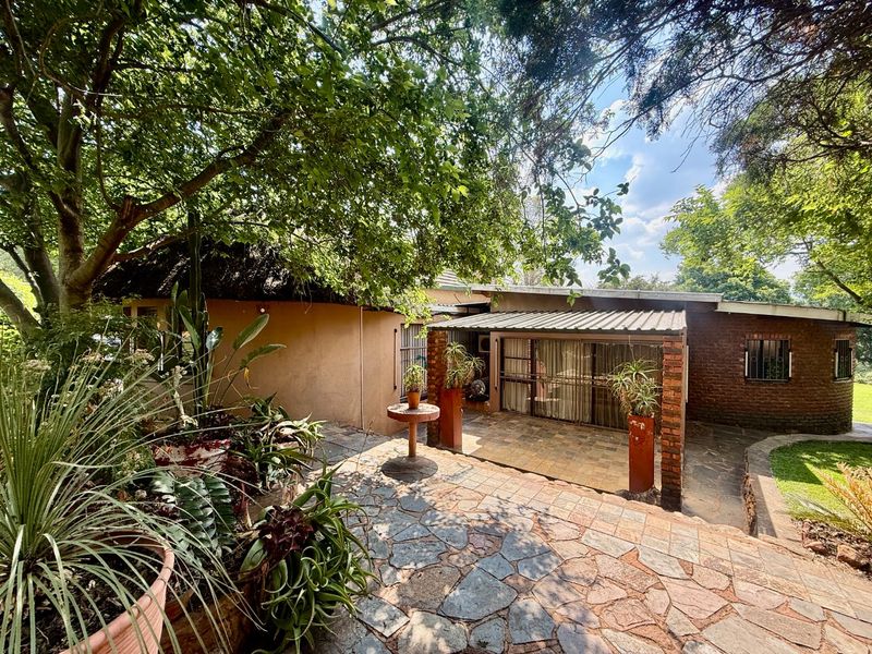 A rear exterior view featuring a stone-paved patio area with a covered section, brick pillars, and mature garden surroundings.